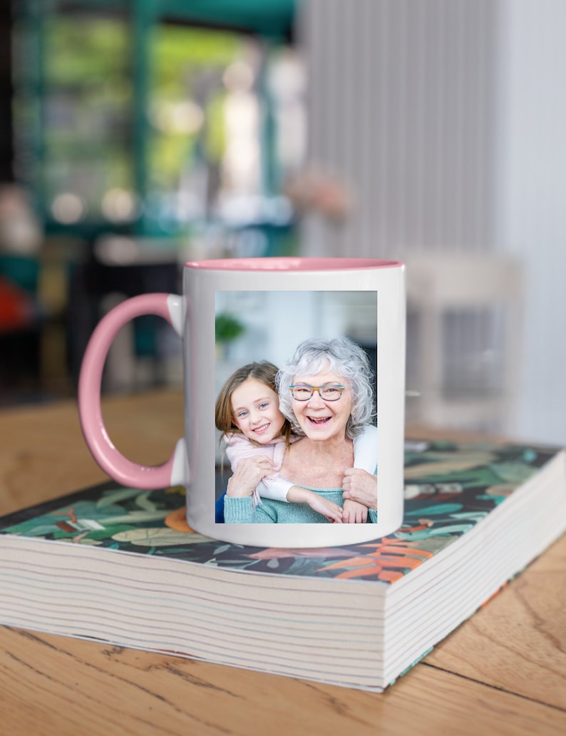 May include: A white ceramic mug with a pink rim and handle. The mug features a photo of an older woman and a young girl smiling at the camera.