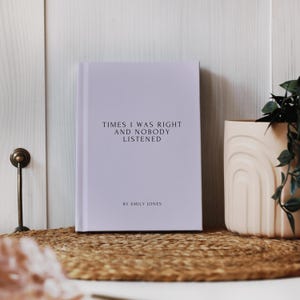 May include: A hardcover book with the title "TIMES I WAS RIGHT AND NOBODY LISTENED" and the author's name "BY EMILY JONES" printed in black. The book is light purple and sits on a woven mat. A beige vase with a plant is to the right.