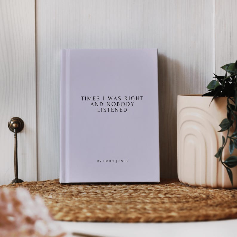May include: A hardcover book with the title "TIMES I WAS RIGHT AND NOBODY LISTENED" and the author's name "BY EMILY JONES" printed in black. The book is light purple and sits on a woven mat. A beige vase with a plant is to the right.