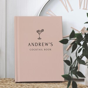 May include: A blush-colored cocktail book with the text "ANDREW'S COCKTAIL BOOK" and a cocktail illustration. The book is hardcover and features a simple, elegant design. The background includes a clock and greenery.