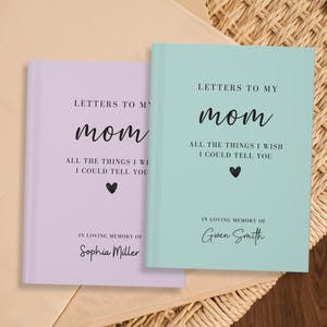 Op de afbeelding: Twee notitieboekjes, een in lavendel en de andere in lichtblauw, met de tekst "Brieven aan mijn moeder" en "Alles wat ik je wil vertellen". De notitieboekjes hebben een zwart hart en de tekst "Ter nagedachtenis aan" gevolgd door een naam.
