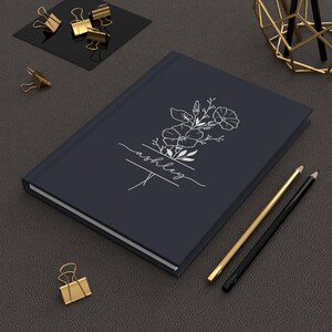 May include: A dark blue journal with a floral design and the name "Ashley" in white script. The journal is closed and has a clean, minimalist aesthetic. Gold and black pencils and gold binder clips are also visible.