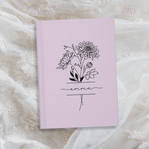 May include: A light purple journal with a floral line art design. The design includes a bouquet of flowers and the name "Emma" in cursive script. The journal is closed and resting on a white lace surface.