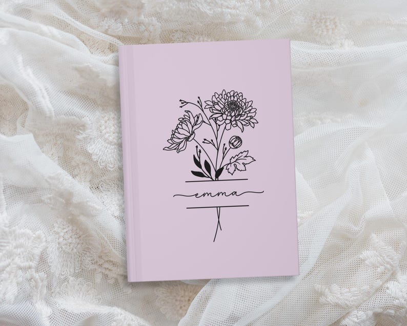 May include: A light purple journal with a floral line art design. The design includes a bouquet of flowers and the name "Emma" in cursive script. The journal is closed and resting on a white lace surface.