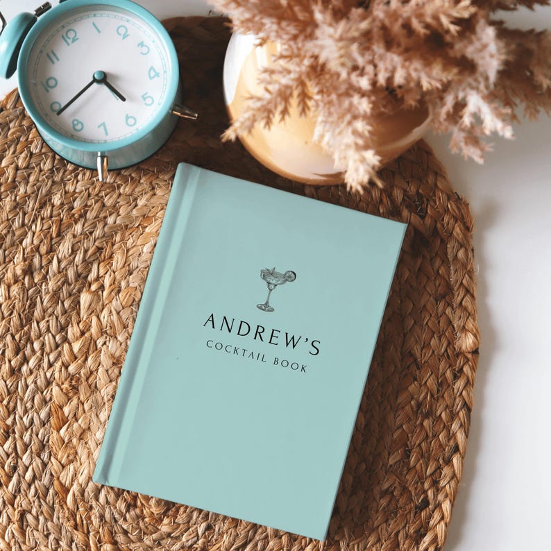 May include: A light blue cocktail book with the text "ANDREW'S COCKTAIL BOOK" and a cocktail glass illustration. A vintage teal alarm clock and a vase with dried flowers are in the background. The book is on a woven mat.