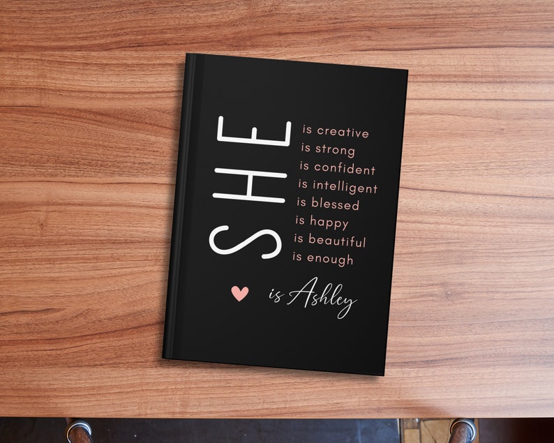 May include: Black journal with white text that reads "SHE is creative is strong is confident is intelligent is blessed is happy is beautiful is enough &hearts; is Ashley".