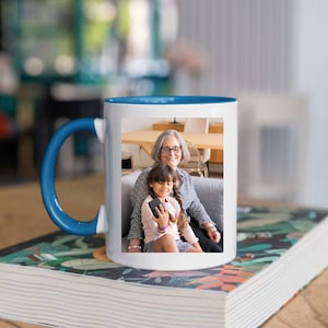 May include: A white ceramic mug with a blue rim and handle. The mug features a photo of an older woman and a young girl smiling at the camera. The photo is printed on the front of the mug.