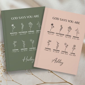 May include: Two hardcover journals, one sage green and one blush pink, with the text "GOD SAYS YOU ARE" and floral illustrations. Each journal features inspirational words like "Beautiful," "Victorious," and "Chosen," along with a personalized name.