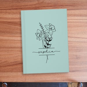 May include: A light green notebook with a floral design and the name "Sophia" in script. The black line art features daffodils and other greenery. The notebook is closed and rests on a wooden surface.