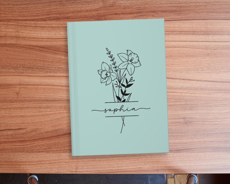 May include: A light green notebook with a floral design and the name "Sophia" in script. The black line art features daffodils and other greenery. The notebook is closed and rests on a wooden surface.