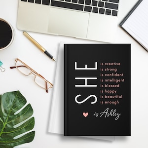 May include: Black journal with the text "SHE is creative, is strong, is confident, is intelligent, is blessed, is happy, is beautiful, is enough &hearts; is Ashley" in white and pink.