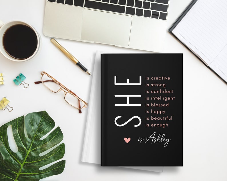 May include: Black journal with the text "SHE is creative, is strong, is confident, is intelligent, is blessed, is happy, is beautiful, is enough &hearts; is Ashley" in white and pink.