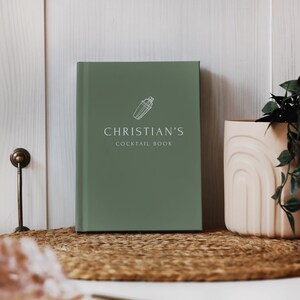 May include: A sage green cocktail book with the text "CHRISTIAN'S COCKTAIL BOOK" in white. A white cocktail shaker icon is above the text. The book is on a woven mat, next to a beige vase with a plant.