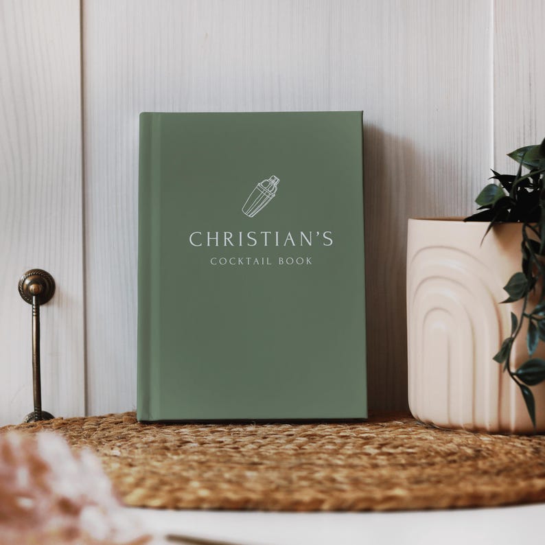 May include: A sage green cocktail book with the text "CHRISTIAN'S COCKTAIL BOOK" in white. A white cocktail shaker icon is above the text. The book is on a woven mat, next to a beige vase with a plant.