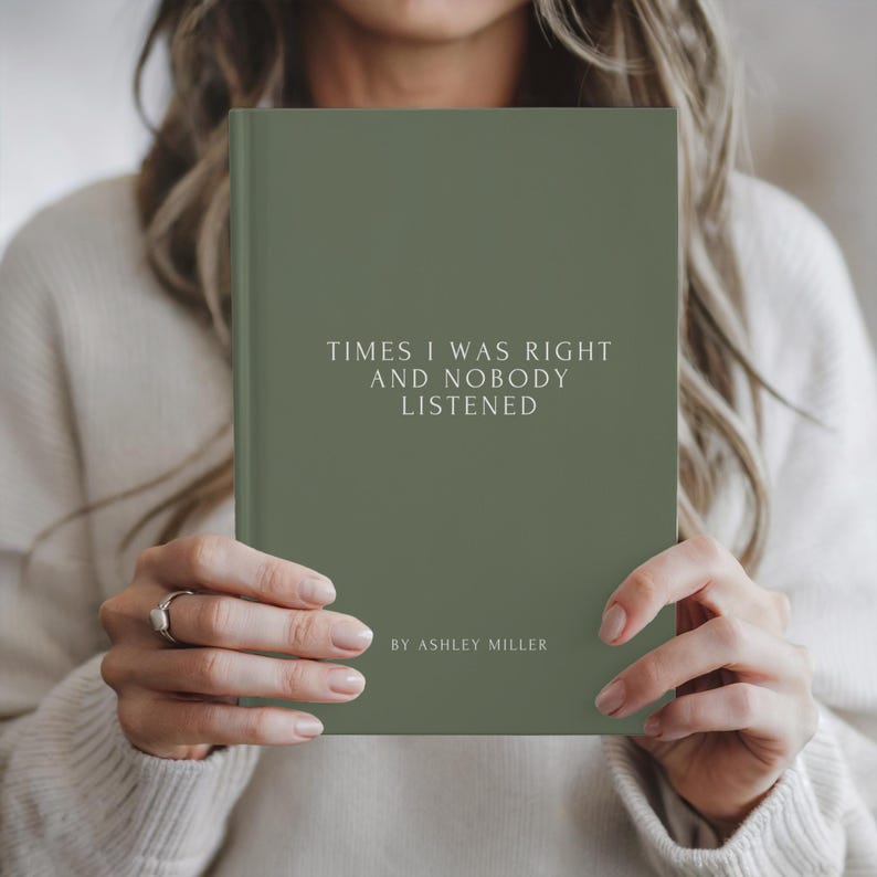 May include: A hardcover book with a sage green cover. The title "TIMES I WAS RIGHT AND NOBODY LISTENED" is printed in white, along with the author's name, "BY ASHLEY MILLER". The book is held by a person wearing a cream-colored sweater.
