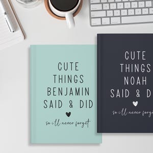 May include: Two blue and green journals with the text "Cute Things [Name] Said & Did so I'll Never Forget" on the front. The journals are on a white surface.