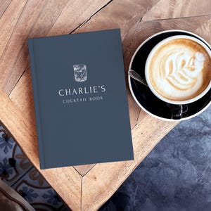 May include: A dark teal cocktail book with the text "Charlie's Cocktail Book" and a sketched image of a cocktail glass. A cup of coffee with latte art sits on a black saucer next to the book on a wooden table.