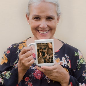 May include: A white ceramic mug featuring a photograph of a family gathered around a table. The photo shows a woman, a girl, and a boy. The woman is holding a plate of food. The mug is being held by a person with grey hair.