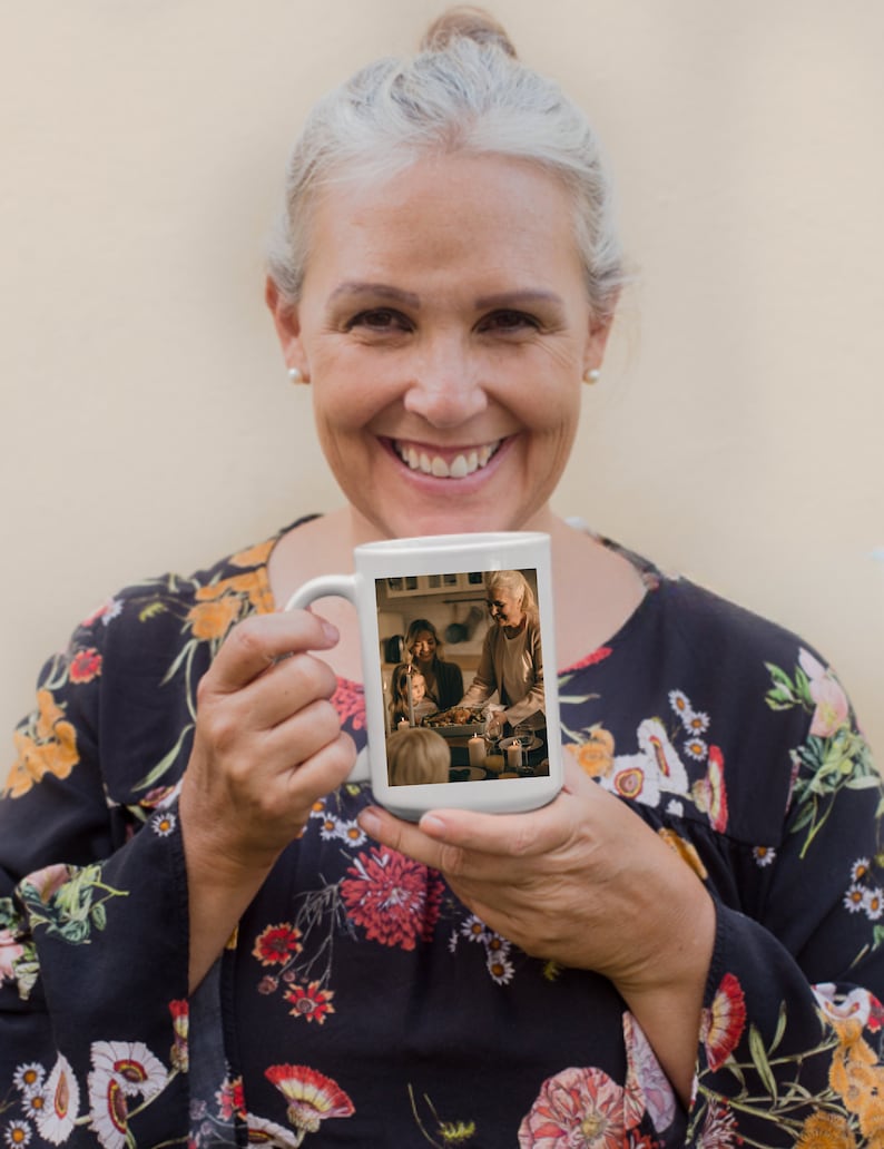 May include: A white ceramic mug featuring a photograph of a family gathered around a table. The photo shows a woman, a girl, and a boy. The woman is holding a plate of food. The mug is being held by a person with grey hair.