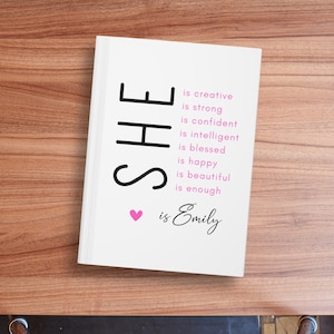 May include: A white book with black text that reads "SHE is creative is strong is confident is intelligent is blessed is happy is beautiful is enough &hearts; is Emily".