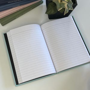 May include: A light blue lined journal with a black spine. The journal is open to a blank page with lined paper. The journal is on a white surface.