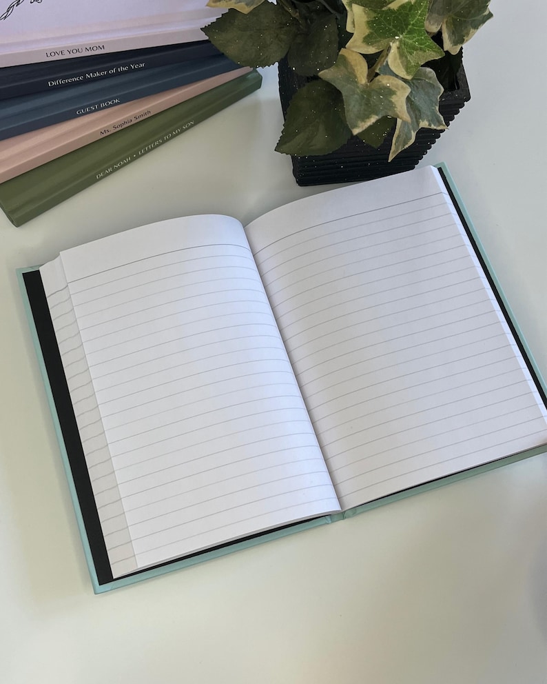 May include: A light blue lined journal with a black spine. The journal is open to a blank page with lined paper. The journal is on a white surface.