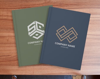 May include: Two closed notebooks with different logos. The notebook on the left has a green cover with a white logo of a geometric shape with the letters "SC" inside. The notebook on the right has a dark blue cover with a gold logo of a geometric shape with a double "S" inside. Both notebooks have the text "COMPANY NAME" and "SLOGAN HERE" printed below their logos.