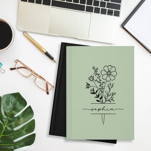 May include: A sage green notebook with a floral design and the name "Sophia" in script. The notebook is next to a black notebook, a laptop, a cup of coffee, glasses, a pen, and a monstera leaf on a white surface.