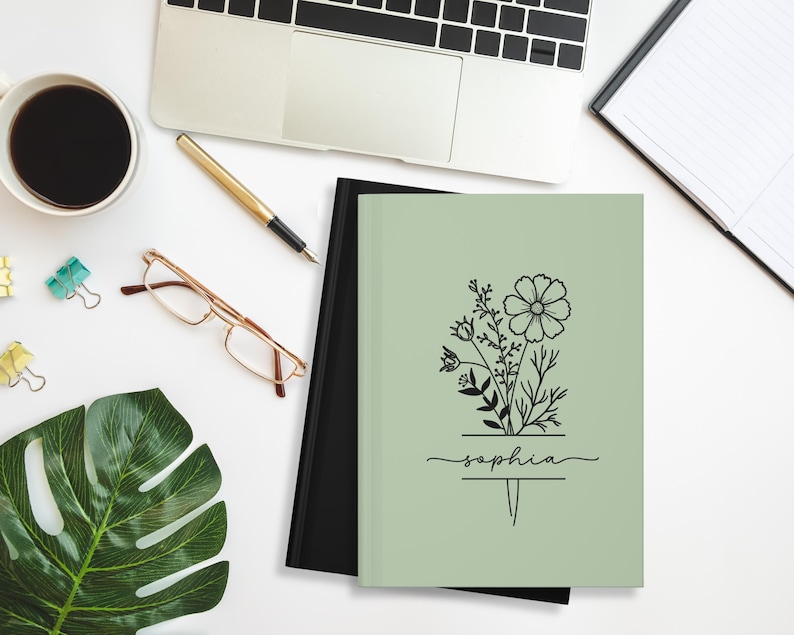 May include: A sage green notebook with a floral design and the name "Sophia" in script. The notebook is next to a black notebook, a laptop, a cup of coffee, glasses, a pen, and a monstera leaf on a white surface.