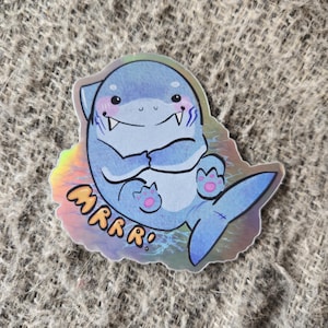 May include: A blue and white holographic sticker of a cartoon shark with pink cheeks and the word "MRRR!" in yellow text.