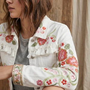 Op de afbeelding: Witte spijkerjas met kleurrijke bloemenborduurwerk. De jas heeft rood, roze en groen geborduurde bloemen met pailletten accenten. Het heeft een franjebies langs de kraag en zoom, en zilveren knoopsluitingen.