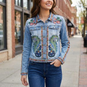Puede incluir: Chaqueta vaquera azul claro con bordados florales y de plumas de pavo real coloridos. La chaqueta tiene cierre de botones, dos bolsillos en el pecho y mangas largas. El bordado incluye tonos de rojo, verde, azul y amarillo.