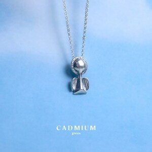 May include: Silver angel pendant necklace with a delicate chain. The pendant is a simple design with a round head and small wings. The necklace is on a light blue background with the text "CADMIUM green" below the pendant.