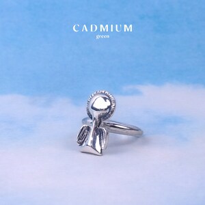 May include: Silver ring with an angel design. The angel has wings and a halo. The ring is on a blue background.