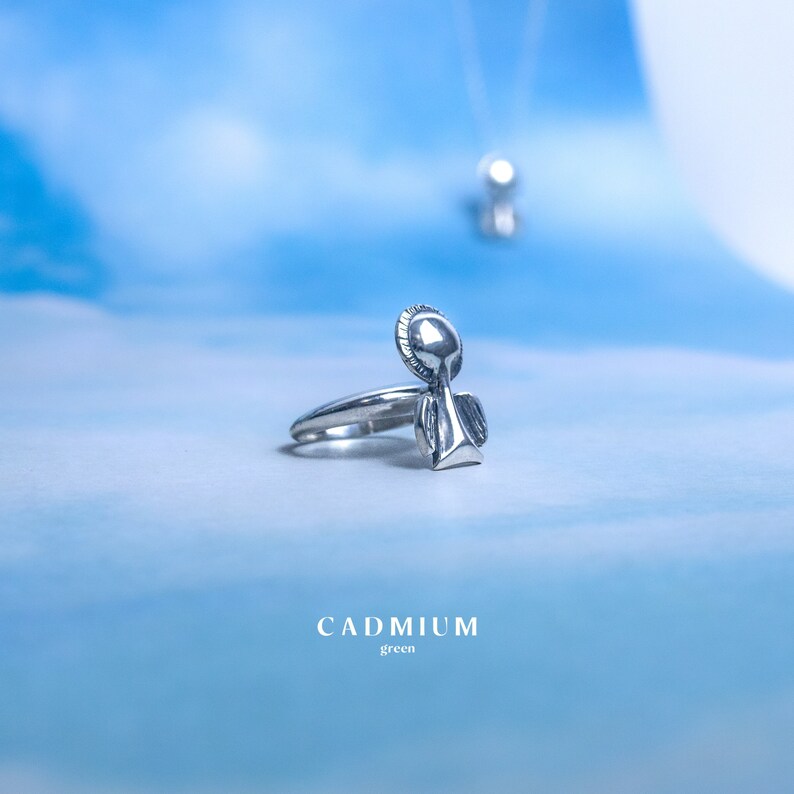 May include: Silver ring with an angel design. The angel has wings and a halo. The ring is on a blue background with the word "CADMIUM" and "green" below it.