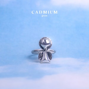 May include: Silver ring with an angel design. The angel has a halo and wings. The ring is on a light blue background.