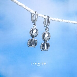 May include: A pair of silver angel hoop earrings hanging from a white rope against a blue sky background. The earrings feature a small angel with wings and a halo. The text "CADMIUM green" is visible at the bottom of the image.