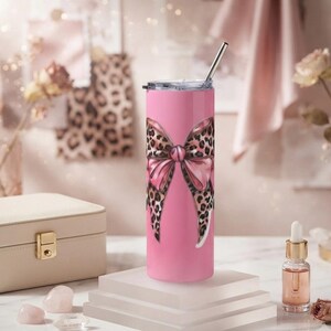 May include: A cylindrical pink tumbler featuring a large, central bow graphic with a brown and black leopard print pattern. The tumbler includes a clear lid and a thin, straight silver metal straw inserted into the top opening of the container.