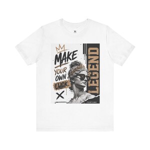May include: A white t-shirt featuring a graphic print of a classical statue head wearing sunglasses. The text reads MAKE YOUR OWN LUCK and LEGEND. The design includes a crown icon, a black and tan color palette, and a distressed artistic style.