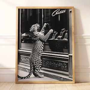 May include: A black and white framed wall art print featuring a leopard standing on its hind legs, looking into a Cartier storefront window display. The display contains jewelry on stands. The frame is made of light-colored wood.