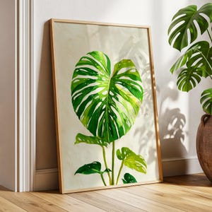 May include: A framed botanical print of a large, green Monstera leaf with white veins. The artwork is set against a neutral background and framed in light wood. The print leans against a wall, with a real Monstera plant in the background.