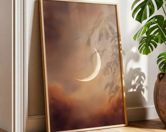 May include: A framed art print featuring a crescent moon in a warm, atmospheric sky. The artwork has a gold frame and is leaning against a wall. The color palette includes shades of brown, orange, and white, creating a serene and celestial theme.