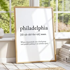 May include: A framed print with the word "philadelphia" in a bold, black font. Below the word is the phonetic pronunciation and the word "noun." The print also includes the text: "Where cheesesteaks are a food group and parallel parking is an extreme sport."