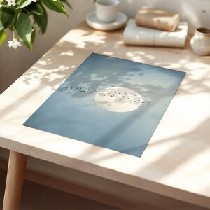 Pu&ograve; includere: Una stampa artistica rettangolare con un paesaggio sereno. L'opera raffigura un grande sole pallido in un cielo azzurro, con uno stormo di uccelli in silhouette. La stampa &egrave; esposta su un tavolo di legno chiaro.