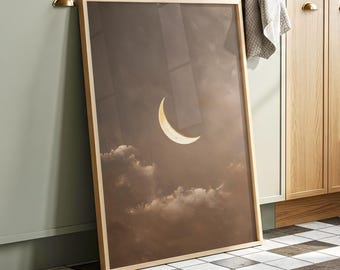 May include: A framed art print featuring a crescent moon in a sepia-toned sky with fluffy clouds. The artwork has a light wooden frame and is leaning against a wall. The overall theme is celestial and atmospheric.