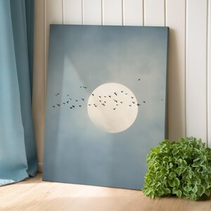 Pu&ograve; includere: Una stampa artistica incorniciata con un grande cerchio giallo pallido, forse il sole o la luna, su uno sfondo blu tenue. Uccelli silhouette sono raffigurati in volo, aggiungendo un senso di movimento. L'opera d'arte &egrave; esposta vicino a una pianta verde.