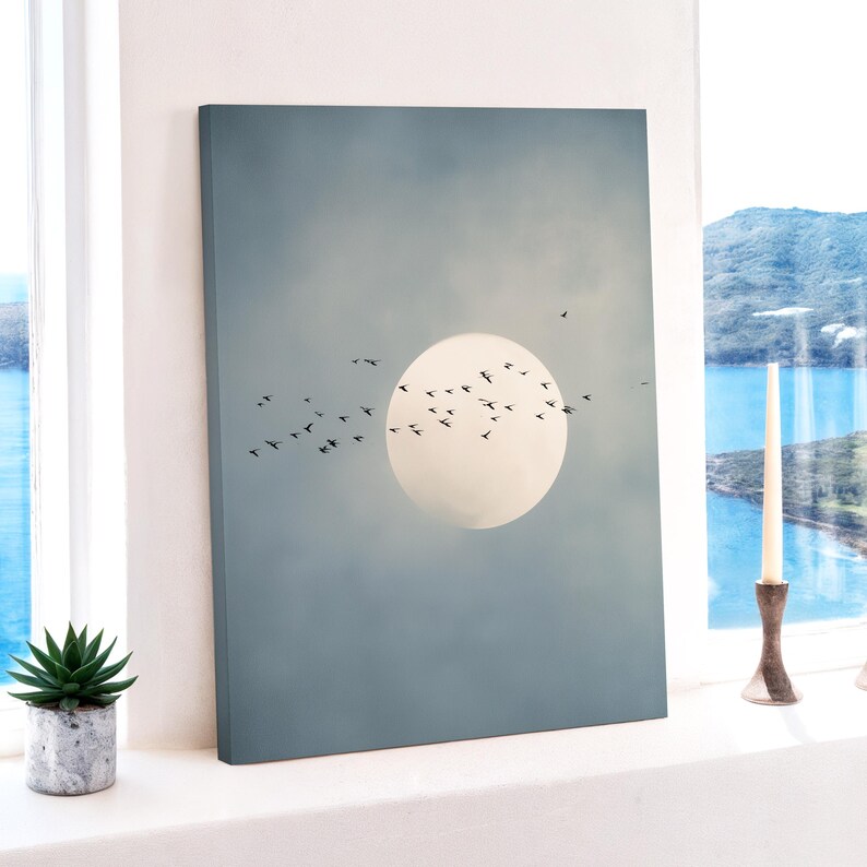 Pu&ograve; includere: Una stampa su tela raffigurante un grande cerchio pallido, simile al sole, su uno sfondo di cielo blu tenue. Uccelli silhouette volano davanti al cerchio. L'opera d'arte &egrave; esposta vicino a una finestra con una piccola pianta succulenta in vaso.