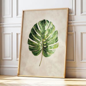 May include: A framed print of a green monstera leaf, showcasing detailed veining and varying shades of green. The leaf is set against a neutral beige backdrop, within a light wooden frame. The artwork evokes a tropical, natural theme.