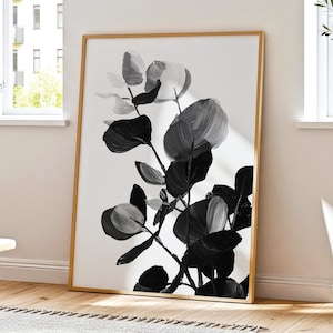 May include: Monochrome botanical print in a gold frame. The artwork presents a stylized illustration of large leaves and stems, using shades of grey and black on a white backdrop. The frame is a warm, natural wood colour.
