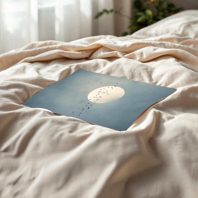 Pu&ograve; includere: Una stampa artistica quadrata con un grande sole luminoso contro un cielo azzurro pallido. Uccelli silhouette volano nel cielo. La stampa &egrave; appoggiata su un letto coperto da un lenzuolo beige.